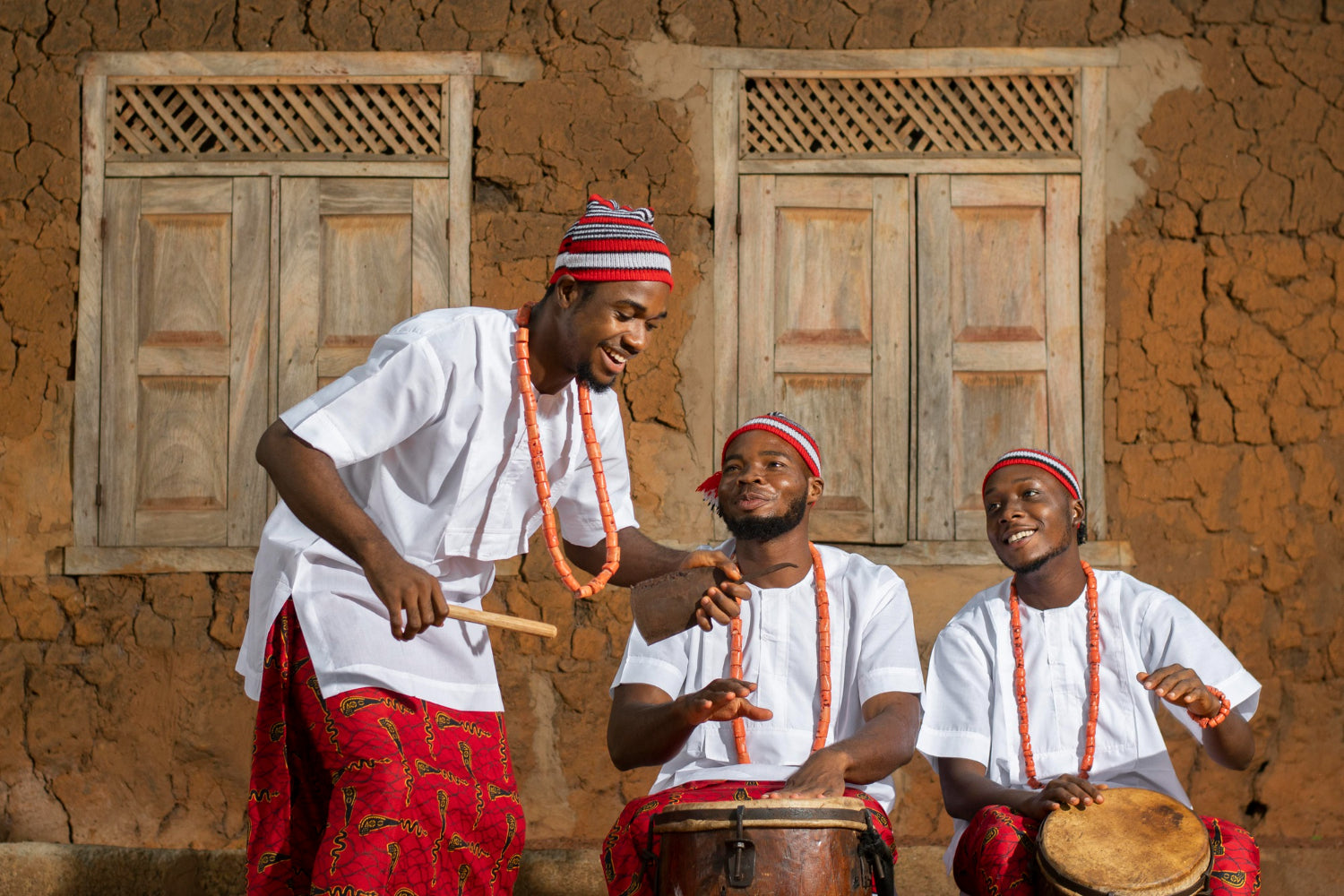 Three men playing drums in front of a traditional building Igbo culture